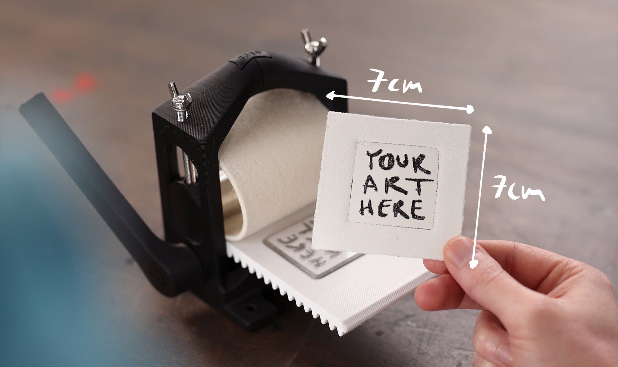 A hand is holding a small 7x7cm sized print into the camera. It shows the words "Your Art here" in black letters. A small black 3D printed printing press is placed in the background.