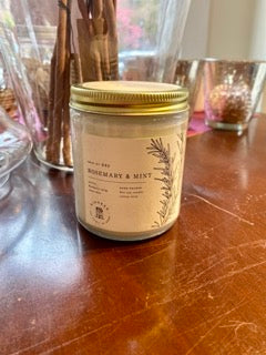 Image of Rosemary & Mint Candle. The candle is in a mason jar with gold screw on top and is sitting on a table. 