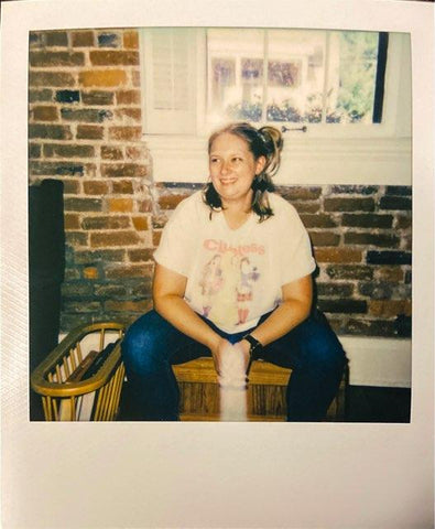Polaroid of Cassie sitting on a chair smiling. Wearing jeans and a white crop T-shirt.