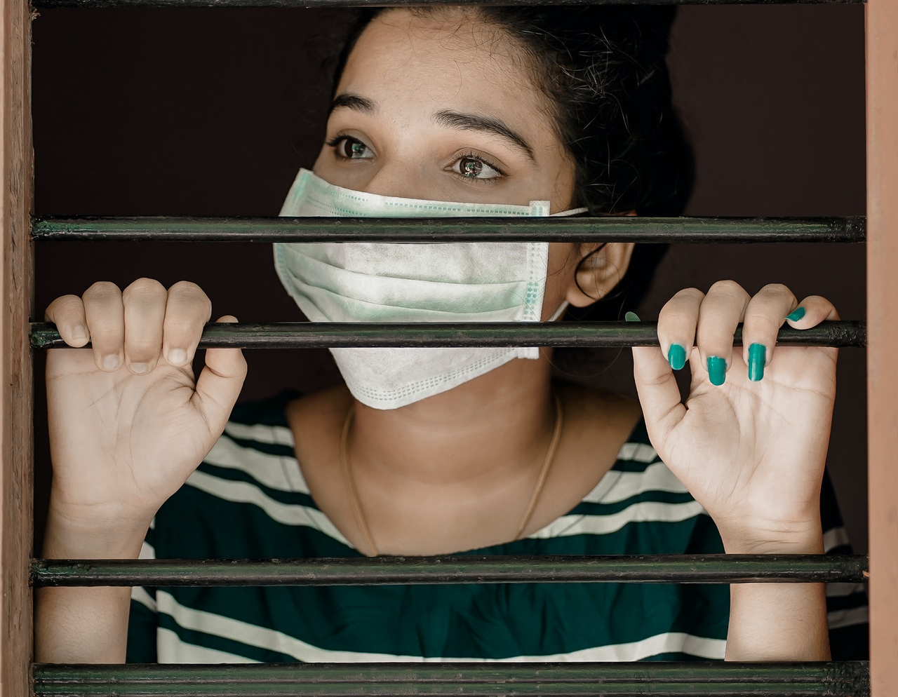 Is The COVID-19 Dilemma Over? A young woman wearing a surgical face mask behind jail bars.