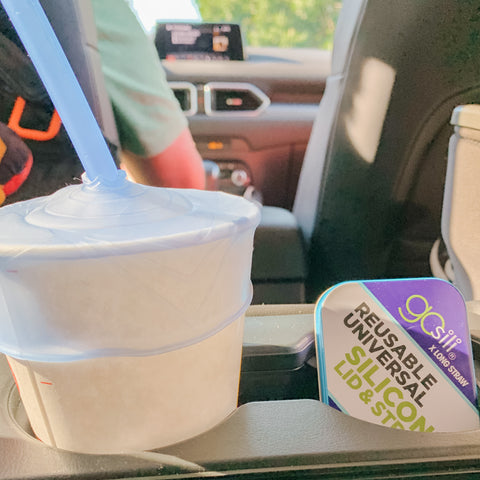 A paper cup with a stretchy silicone lid and a silucone straw in it. The cup sits in a car with the driver on the background. 