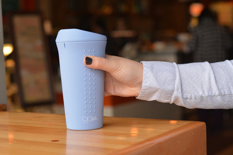 A hand reaching out to grab a blue silicone cup with a coffee shop as background.