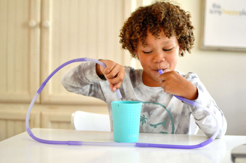 A kid sitting on a counter with a cup and four connected silicone straws. He's smiling as he blows on one end of the straw and water comes out on the other end.