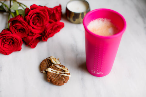 A reusable silicone cup filled with a vegan pumpkin latte. Next to it there are two cookies, a candle, and a bouquet of roses
