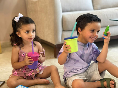 Two kids sitting on the floor holding silicone reusable cups with straws 