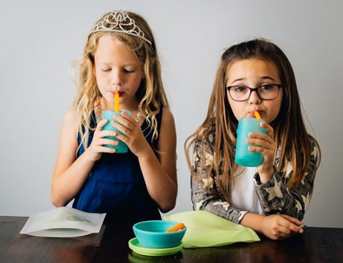 Two school-age girls sip water from a reusable silicone tumbler using a silicone straw. There's a table in front of them with a set of silicone snack bags and a silicone bowl with lid