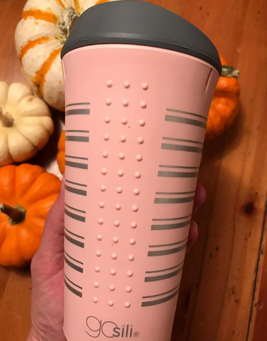 A pink reusable silicone cup with a silver stripes is held by a hand. On the background you can see orange and white pumpkins. 