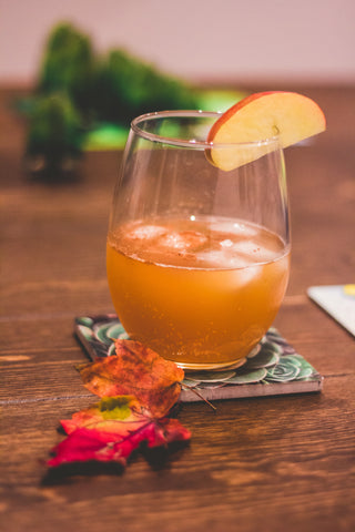 A clear glass with iced apple cider in it. It has an apple slice as garnish and is sitting on a coaster with autumn leaves by its side. 
