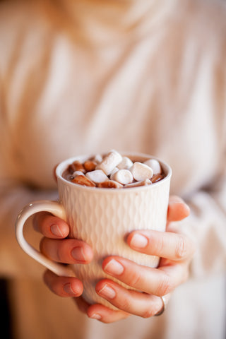 A set of woman's hands is holding a white cup with hot chocolate with marshmallows in it.