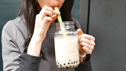a woman holding a glass jar filled with boba tea. She's sipping at her boba tea using a wide reusable silicone straw.