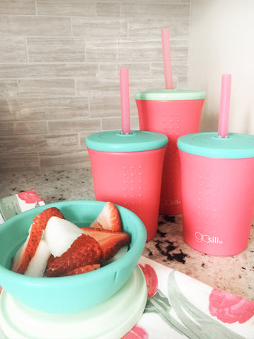 Three reusable silicone tumblers and a reusable silicone bowl with lid filled with strawberries and yogurt sit at a kitchen counter