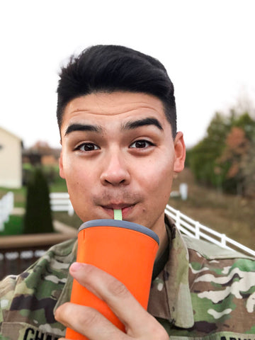 Guy on camouflaged jacket looking directly at the camera is sipping from an orange silicone cup with a grey top and a white straw.