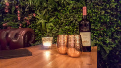 A picture of 2 CLINQ Hammered Stemless Copper Glasses on a wooden table with a bottle of red wine to the right and a candle to the left.