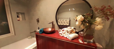 A front on image of a hammered, copper basin made by Empire Copper. The basin sits upon a red wood bathroom counter. There is a large round mirror behind it and loads of flowers on the right hand-side of the counter.