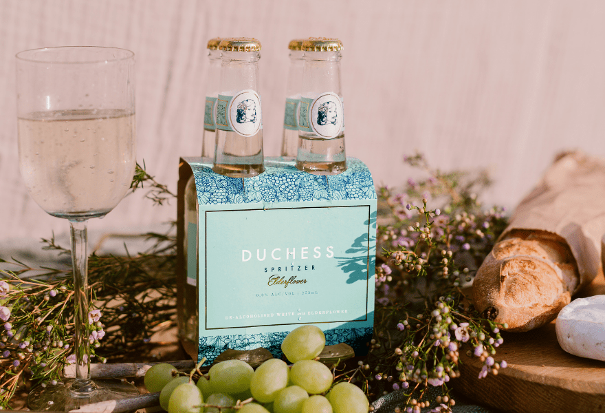 A pack of 4 The Duchess Alcohol-Free Wine Spritzer Elderflower next to a glass of low calorie non-alcoholic wine, grapes and bread