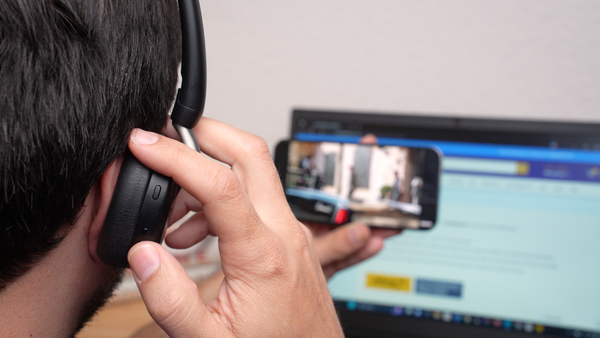 man wearing an Evolve2 65 Flex headset holding a mobile phone while on a live video call with a computer in the background