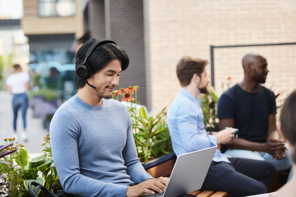 Man sitting in the foreground in an office wearing a Jabra Evolve2 75 headset, with coworkers talking in the background