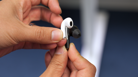 close up view of man holding one Apple Airpod earbud and Poly Voyager Freee 60 earbud side by side