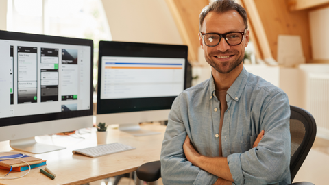 picture of a smiling IT professional sitting in front of two large computer monitors with a smile, and arms folded.