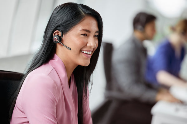 Woman sitting at an office desk wearing a Jabra Engage 55 earpiece while smiling