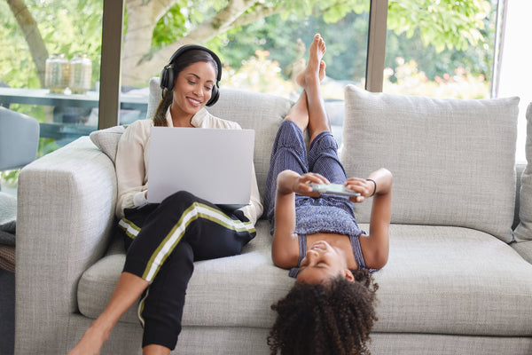 A Woman sitting on a couch with a laptop and wearing a Jabra Evolve2 75 headset, and a child jumping on the couch next to her