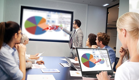 People participating in a video meeting with a presenter in front standing up, others sitting around a conference table with one person sharing video content from a  laptop computer