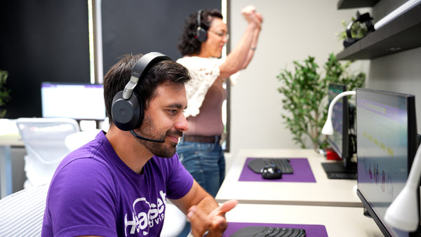 man in foreground weairng jabra evolve2 85, and woman in background standing up  in  front of computer with arms  raised, making noise on a call.