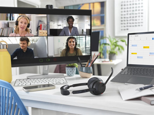 desk with monitor showing 4 people on a video call with headset resting on desktop, and within sight is a laptop, and a mobile phone