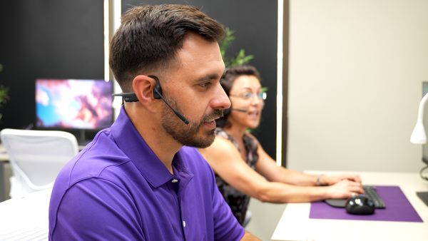 Man in foreground at office desk wearing an Opencomm2 UC headset, with coworker in background