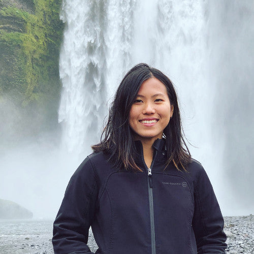 a dark haired woman in a black jacket smiling in front of a waterfall
