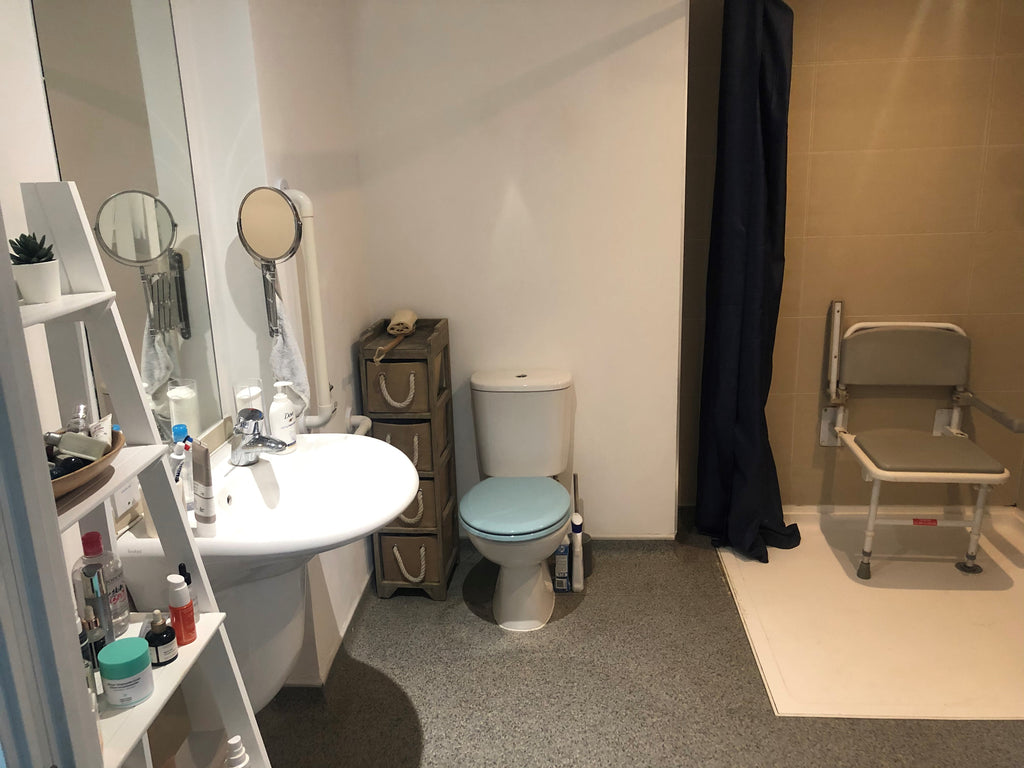 Sophie Morgan's bathroom before. Tired looking room with grey flooring and white shower tray, white walls, light blue toilet seat, padded grey shower chair with beige tiles behind and dark blue shower curtain