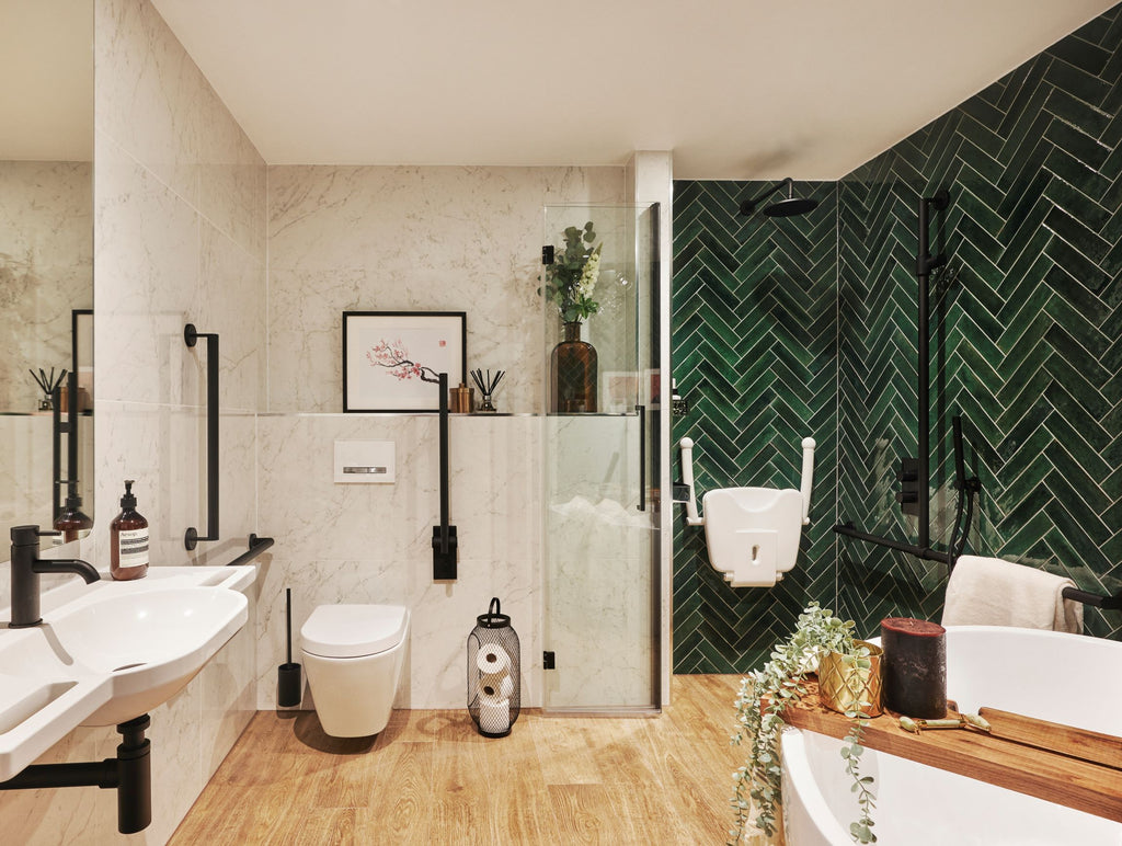 Sophie's new bathroom. Toilet area is white marble tiled, shower area is deep mottled green subway tiles laid in a herringbone pattern. Wood-effect floor tiles throughout and matt black grab rails. Artwork, flowers and candles sit on a built-in shelf behind the toilet. The edge of a large curved bath in the foreground has a wooden bath board across it, decorated with plants and bath salts. A white shower seat (in folded up position) is in the shower area behind a single folding glass screen.