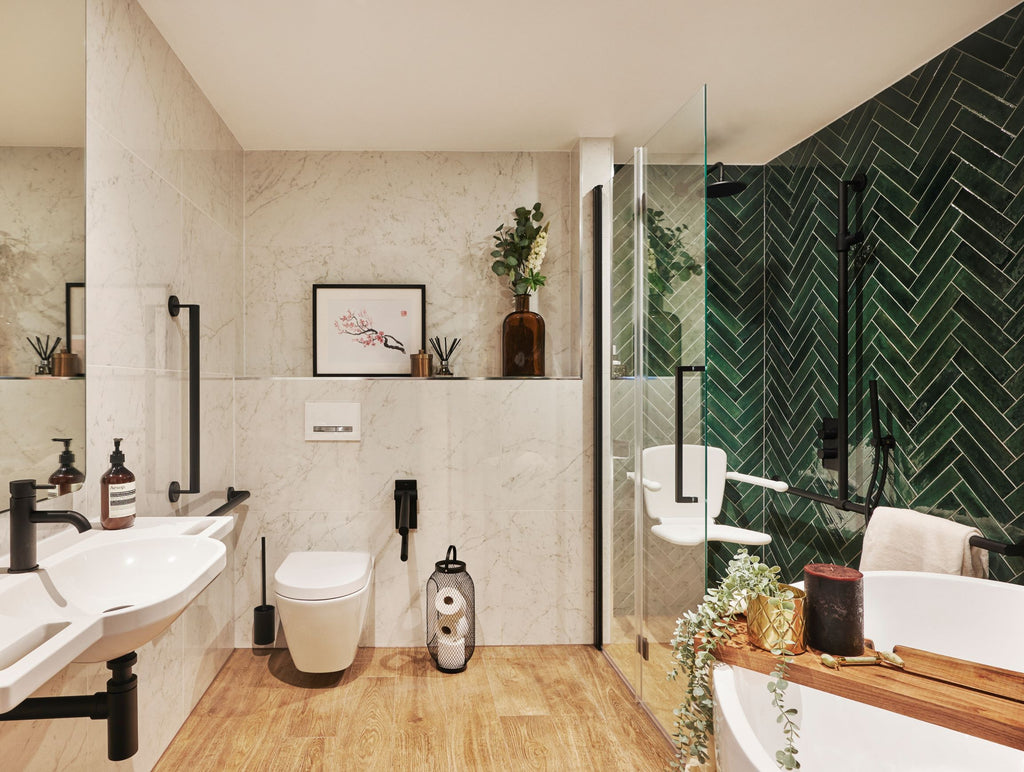 Sophie's new bathroom. Toilet area is white marble tiled, shower area is deep mottled green subway tiles laid in a herringbone pattern. Wood-effect floor tiles throughout and matt black grab rails. Artwork, flowers and candles sit on a built-in shelf behind the toilet. The edge of a large curved bath in the foreground has a wooden bath board across it, decorated with plants and bath salts. A white shower seat is in the shower area behind a single folding glass screen.