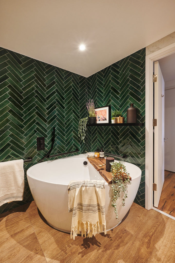 Corner view of Sophie's new bathroom, focused on curved floor standing corner bath. Surrounded by rich mottled green subway style tiles in a herringbone pattern. Wood-effect floor tiles on the floor and a black shelf full of plants and artwork just above the bath. A wooden bath board sits across the bath, decorated with plants, towels and bath salts.  Matt black grab rails subtly blend into the green tile around the bath.