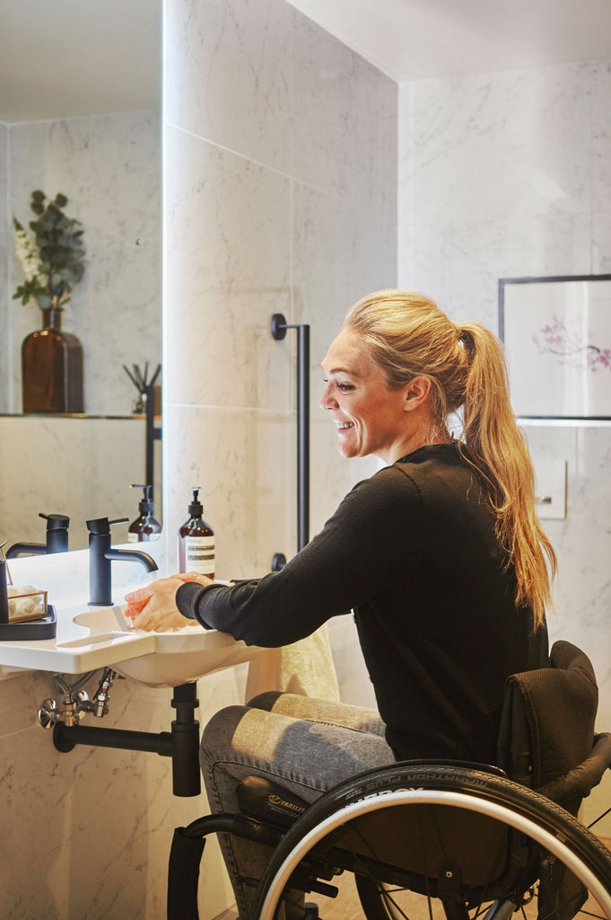 Sophie looking happy, washing her hands over the wall hung basin in her new bathroom, seated in a wheelchair, with illuminated mirror giving off a soft halo of light. 