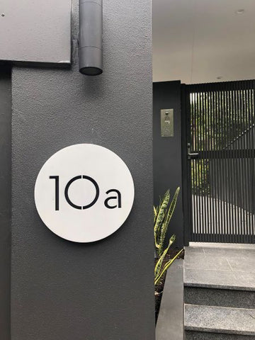Modern Round, brushed aluminum plaques '10a'. On dark grey exterior wall with black gate in the background.