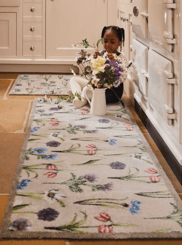 A young girl sat with a vase of flowers on a floral kitchen runner