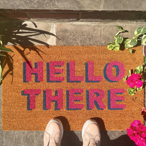 A pair of feet stood on a coir outdoor mat saying hello there in pink writing