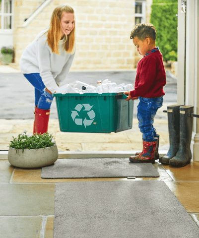 Children carrying a recycling box over a grey doormat