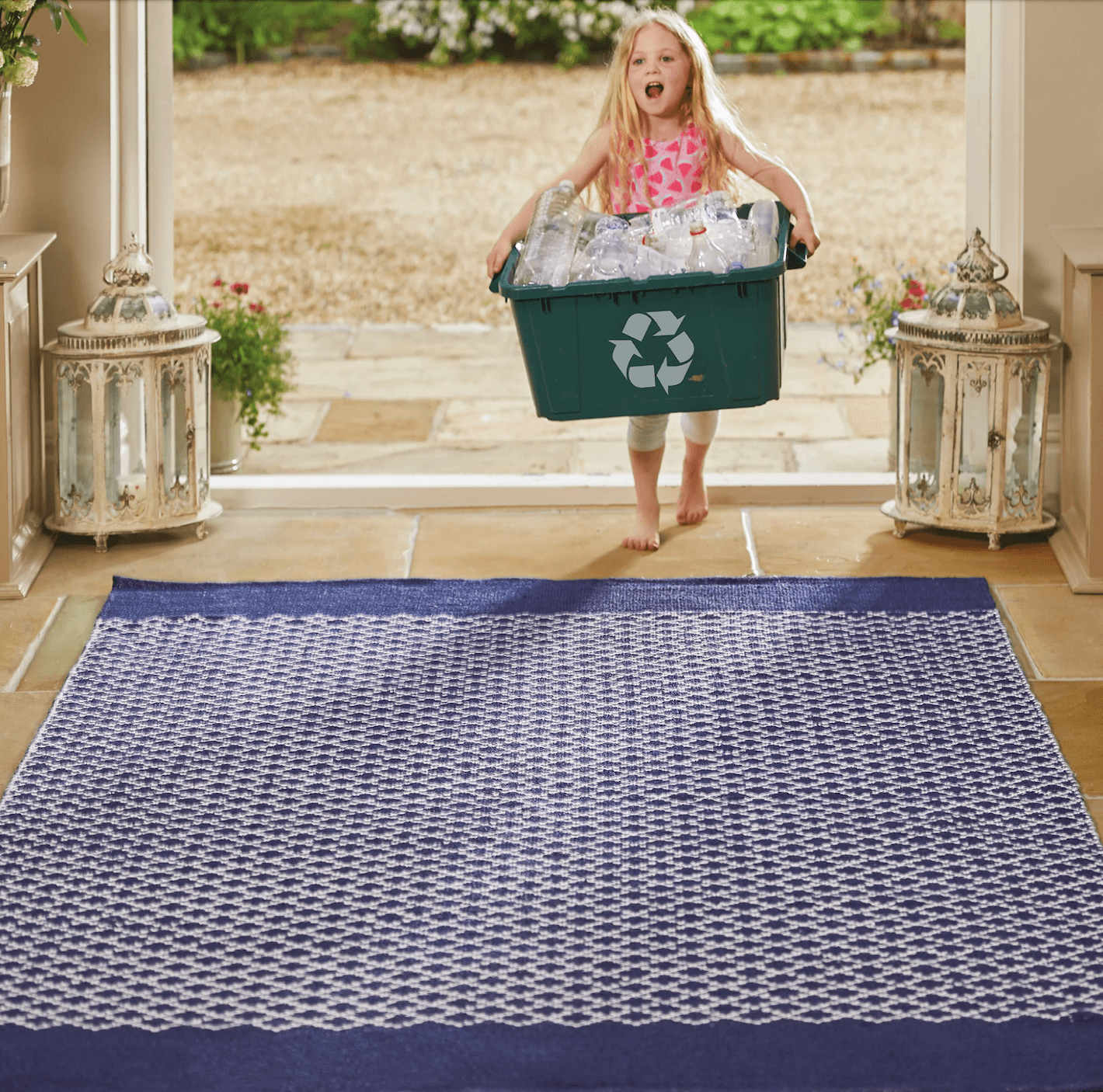A young girl carrying a box over plastic bottles over a blue woven rug