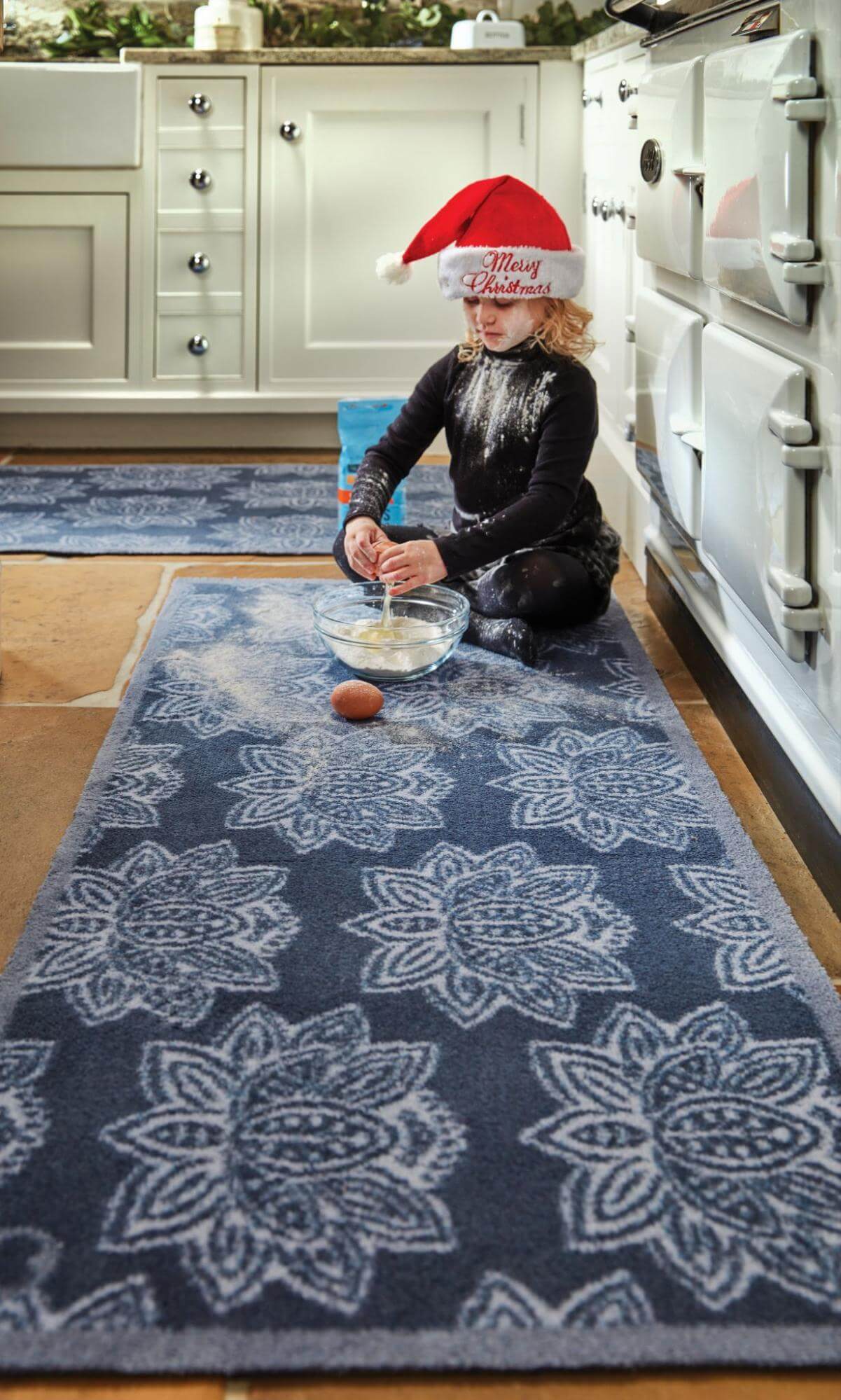 A young girl sat on a kitchen runner cracking an egg into a bowl wearing a santa hat