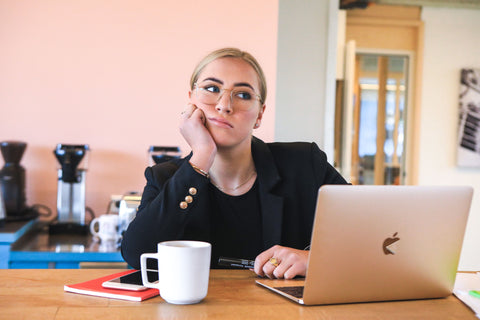 A blonde woman sitting at a desk, resting her chin in her hand, deep in thought in front of her MacBook