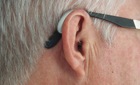 An elderly person with grey hair and glasses is wearing one hearing aid on his ear