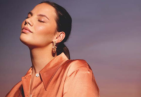 Model with hair in ponytail and in orange shirt poses with her eyes closed against a sunset background.