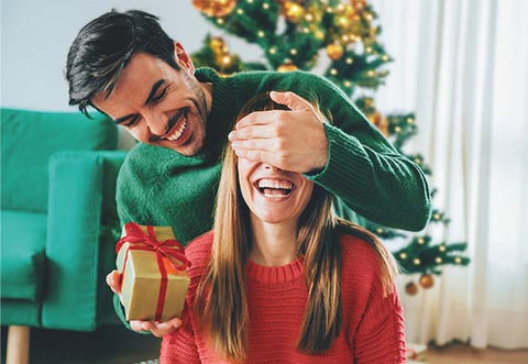 Man sits behind woman with his hand covering her eyes and a gift box in the other hand.