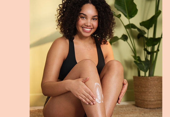 Summer skincare - model applies cream to her legs with plant in the background.