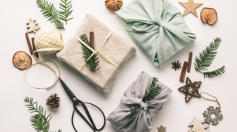 A photo of cutely decorated gifts, tied in cloth instead of paper, with various pieces of greenery and natural decor like wood chips and cinnamon sticks scattered around.