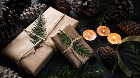 A moody photo of two gifts wrapped in brown paper, tied with string and some pieces of fir tree as decoration, beside some tea light candles.