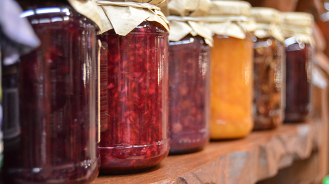 Six jars of homemade jelly on a shelf, the lids are covered with a small piece of fabric and tied with string.