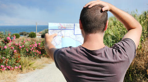 Person in a t-shirt facing away from the camera, holding a map in one hand and their other hand on their head, as if confused.